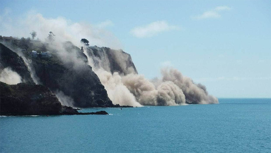 Steep rock slope failure in New Zealand. Clouds of dust rise up from cliffs descending into ocean.