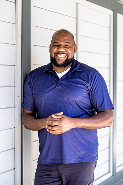 UW Tacoma alumnus Michael Jordan. Jordan is wearing a purple shirt and blue jeans. He is smiling and his right shoulder is leaning against a white wall.