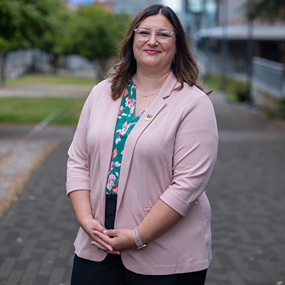 SIAS Dean Natalie Eschenbaum stands on the Prairie Line Trail on campus. She has her arms crossed in front of her by her waist. She is wearing a pink sweater with a floral print shirt underneath. Eschenbaum has long brown hair and wears glasses.