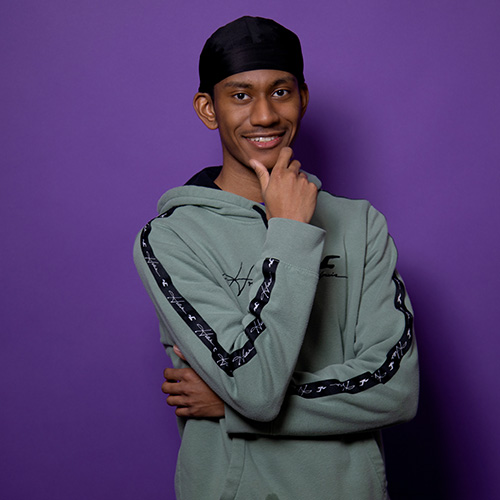 Headshot of UW Tacoma junior Christian James. James is wearing a grey hoodie with black stripes on the sleeves. He has a black cap on his head. James' right hand rests underneath his chin. He stands against a purple background.