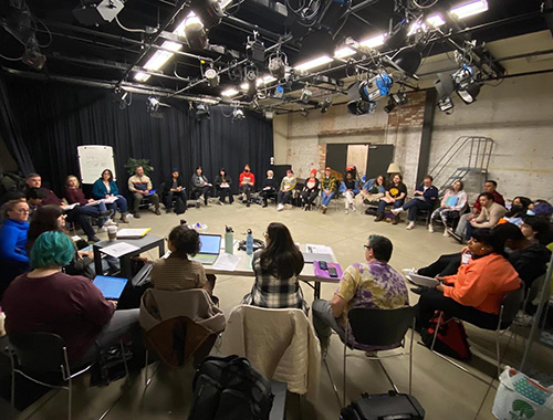 The cast of the musical Rock of Ages sits in a circle inside the Cherry Parkes building on campus. The group are sitting in chairs. 