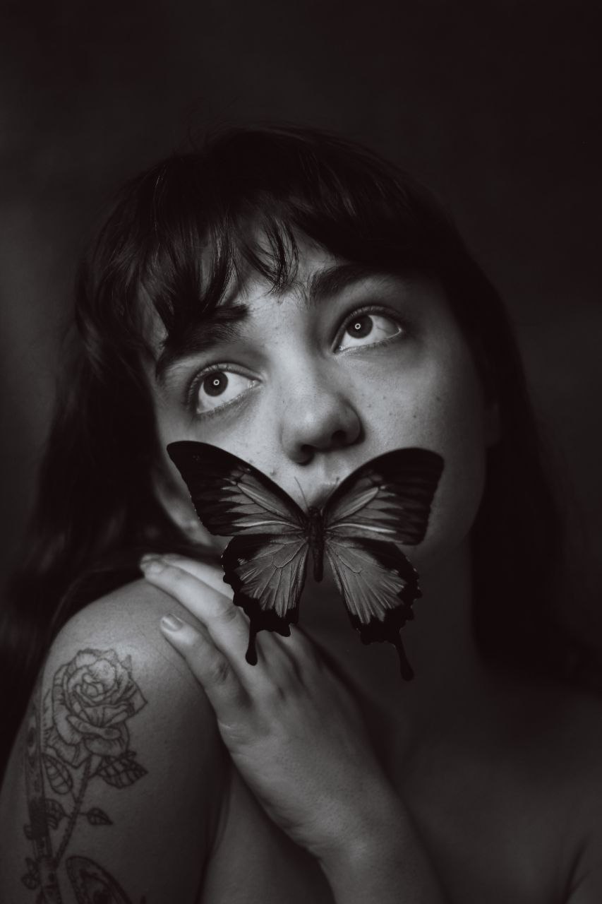A close up portrait of a woman cropped to show her from the shoulders up. Her left hand rests on her right shoulder, which is raised. She is leaning her head toward her hand as she looks to the upper left of the frame. A butterfly obscures her mouth, chin and right cheek.