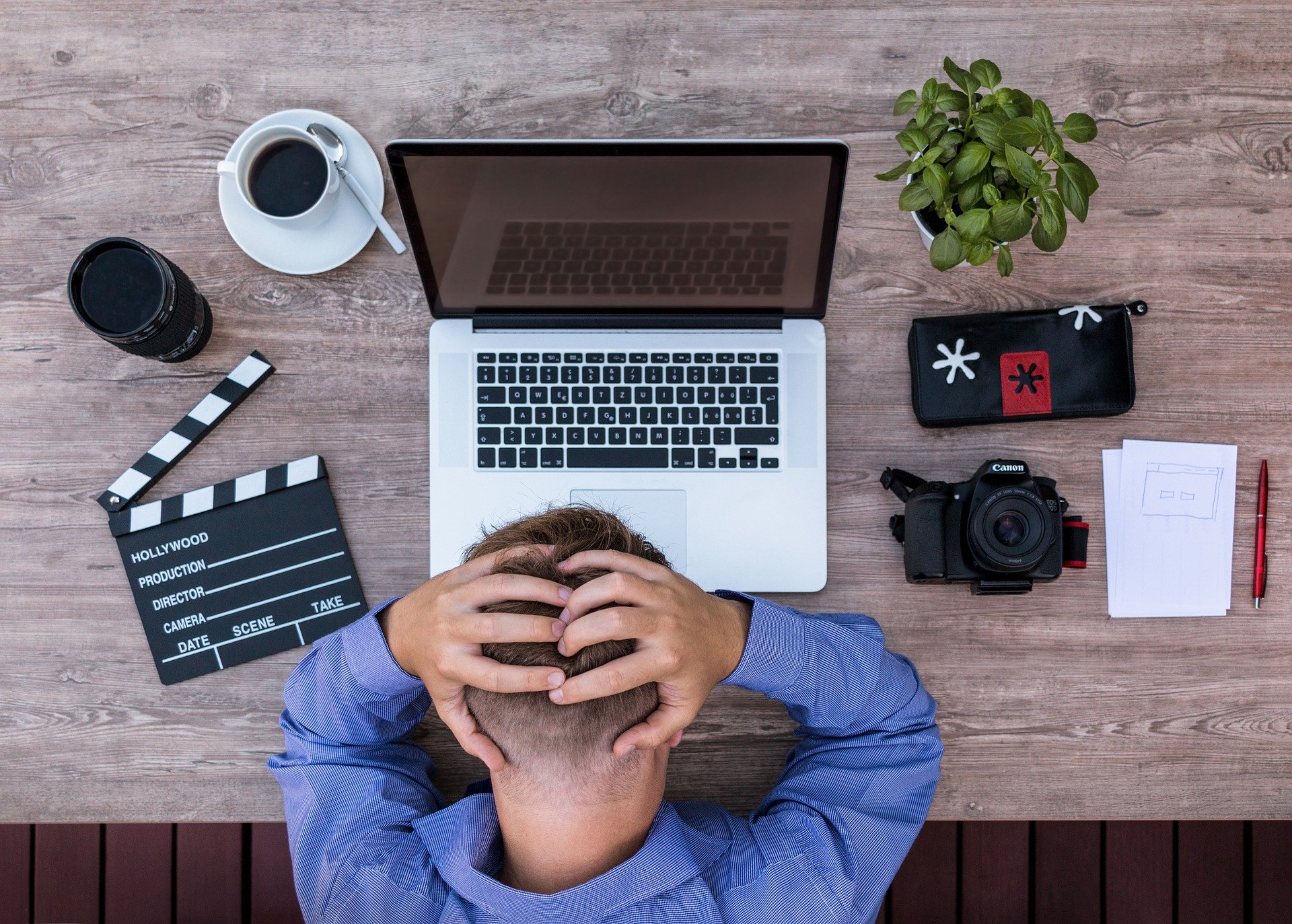 Man with hands on head looking at desk with computer, camera and coffee