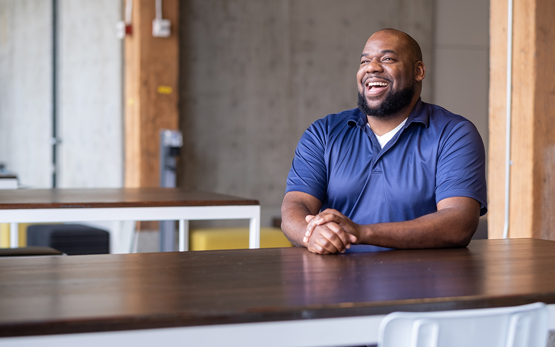 UW Tacoma alumnus Michael Jordan sits at a table. The table has a brown top and white sides. Jordan is wearing a purple shirt and is smiling. His hands are folded and his arms rest on the table top.