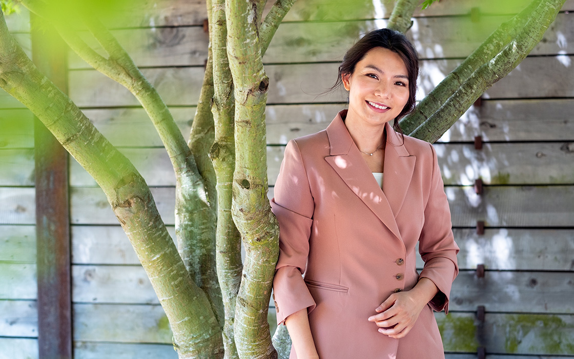 UW Tacoma faulty member Sunny Chieng stands underneath a tree. She has long black hair and is wearing a pink jacket. There is a wooden retaining wall in the background. Leaves from the tree can be seen in the front of the photo.