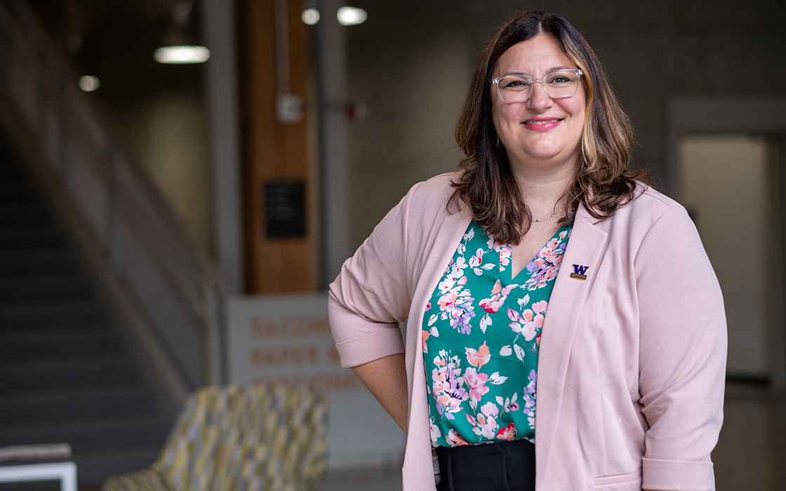 SIAS Dean Natalie Eschenbaum stands with her right hand on her hip. She is inside the Tacoma Paper Stationary building on campus. Eschenbaum is wearing a pink sweater with a floral print shirt underneath. She has long brown hair and wears glasses.