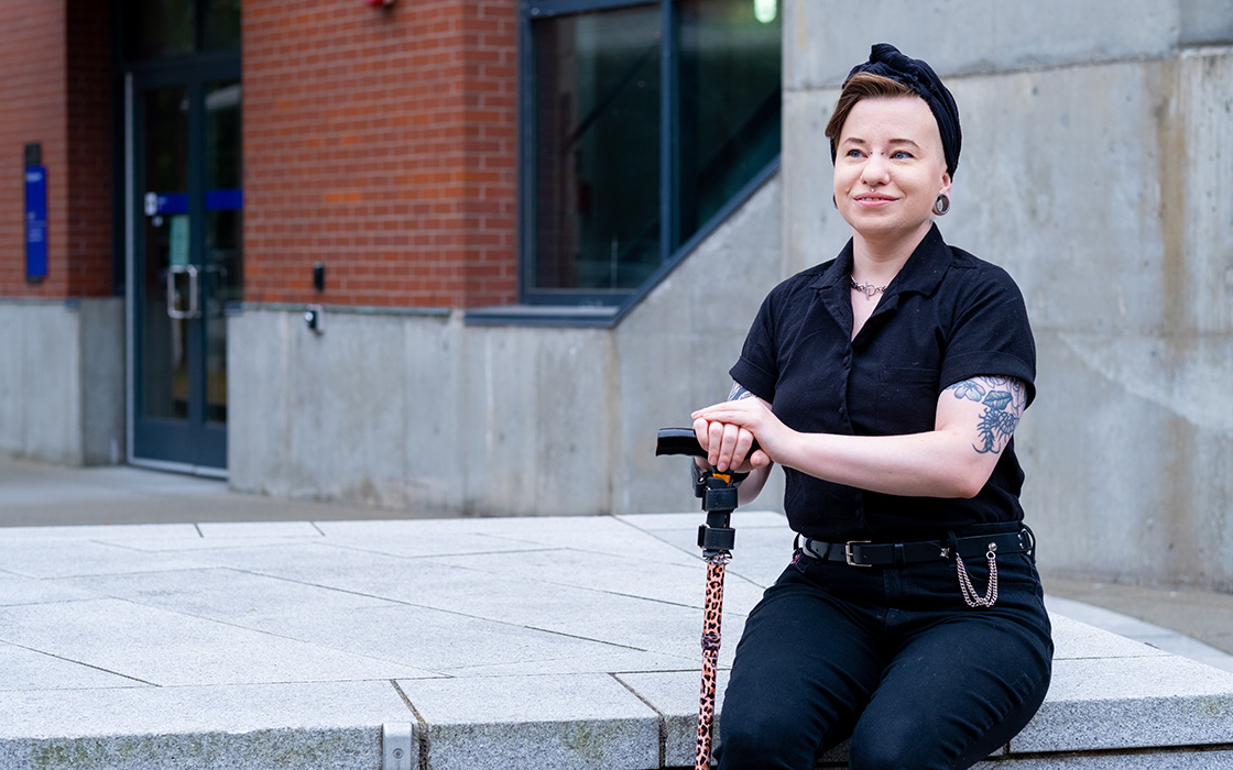 Vern Harner sits on a concrete bench. They are wearing a dark shirt, dark pants and a dark hat. Harner is holding a walking cane. They have a visible tattoo on their left arm and a piercing in their nose.