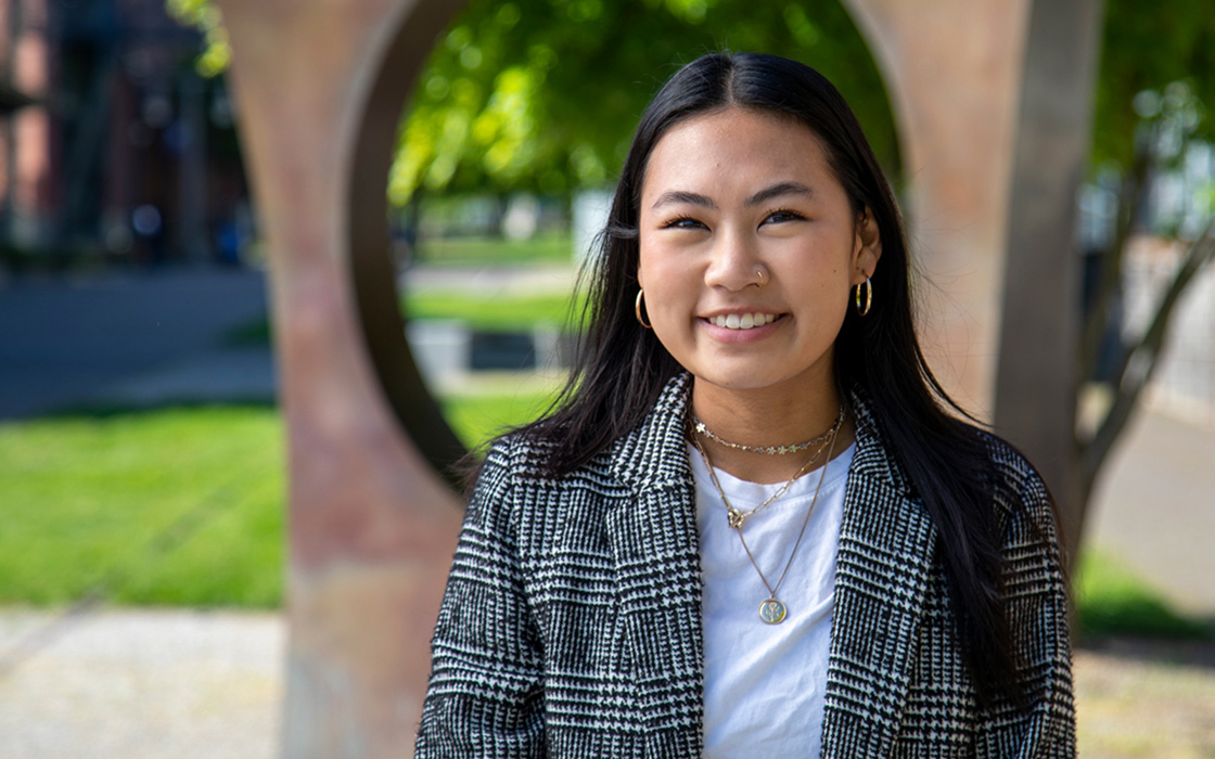 UW Tacoma alumna Teiya Shimomura stands in front of the Maru sculpture on campus. Maru is a bronze, square with a hole in the center. Teiya has long dark hair and is wearing a plaid coat with a white shirt underneath.