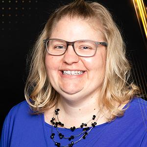 Headshot of staff member BrieAnna Bales. Bales is smiling and wearing glasses. She has medium length dirty blonde hair. She is wearing a purple blouse.