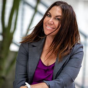 UW Tacoma academic advisor Isabella Webb sits with her arms crossed. She has long, dark hair and is wearing a grey blazer with a purple blouse underneath. In the background is an out of focus tree.