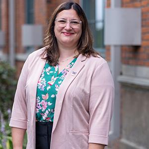 SIAS Dean Natalie Eschenbaum stands in front of the library on campus. She is wearing a pink sweater with a floral print shirt underneath. Eschenbaum has long brown hair and wears glasses.