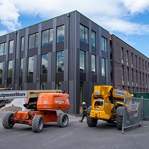 Exterior photo of Milgard Hall. In the front of the building are construction vehicles. One is orange the other is yellow. Milgard Hall is in the background. It's a three story grey and red brick building.