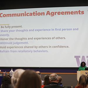 UW Tacoma Vice Chancellor for Equity & Inclusion Elavie Ndura stands on a stage. Behind her is a white screen with a community agreement written on it. The agreements detail the behavior expected of people in the audience.