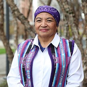 Ada McDaniel sits on a bench. There are trees behind her. McDaniel is wearing a purple headwrap with designs woven into the fabric. She is also wearing a purple vest with designs woven into the fabric. McDaniel is wearing a white undershirt.