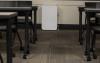 White, rectangular floor-standing air purifier against a wall, seen through an aisle of classroom desks and chairs.