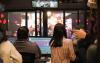 A group of people stand behind a window and look out over the stage at the Tacoma Little Theatre. The people are audio engineers for the show and are checking sound levels.