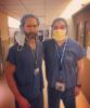 Dr. Michael Mulligan (left) stands next to UW Tacoma student Lucas Bjorkheim. Both men are wearing blue medical scrubs. Mulligan has his hair in cap and his mask off. Bjorkheim also has his hair in a cap and is wearing his mask. They are in the hallway of a hospital.